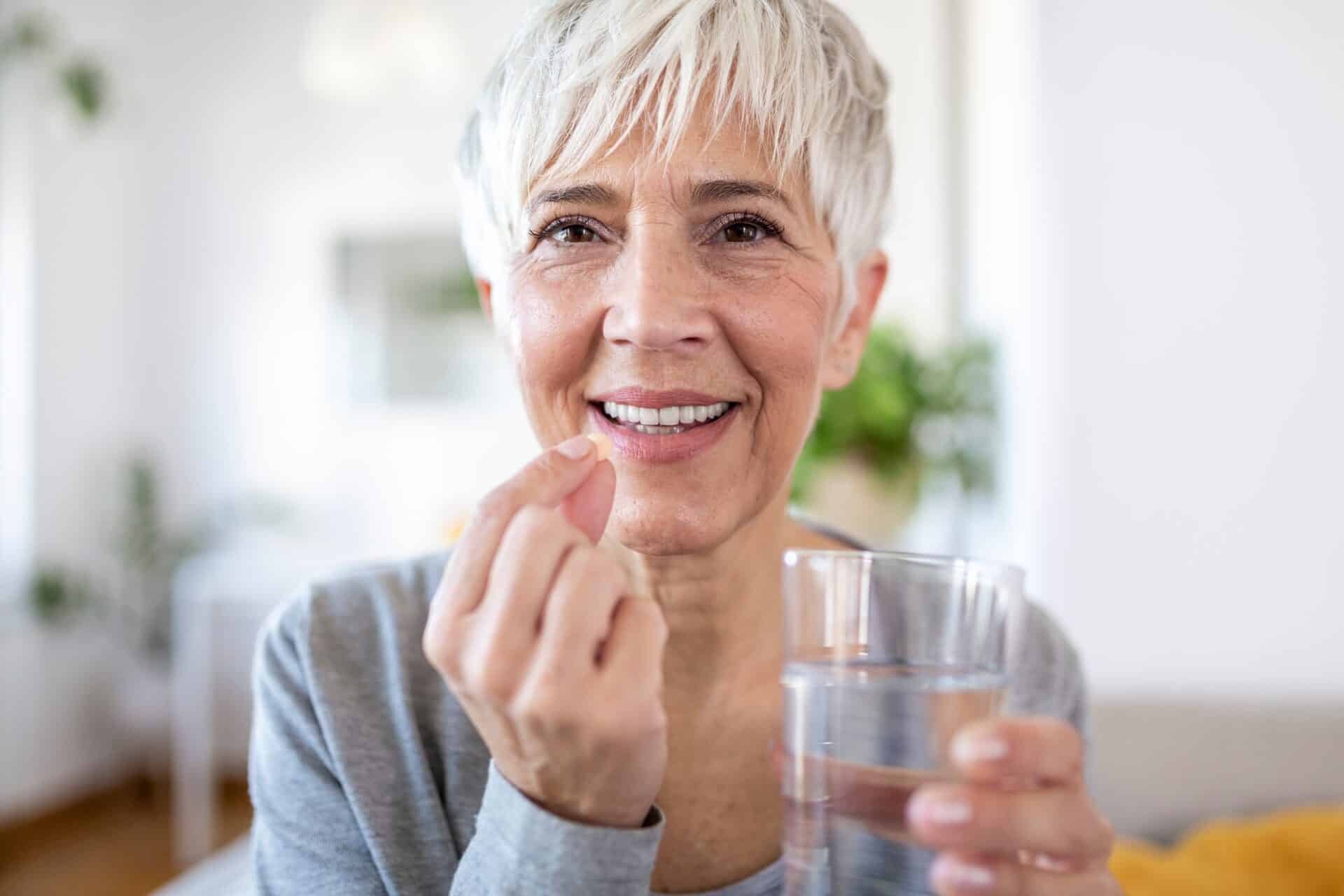 Smiling happy healthy middle aged 50s woman holding glass and pill